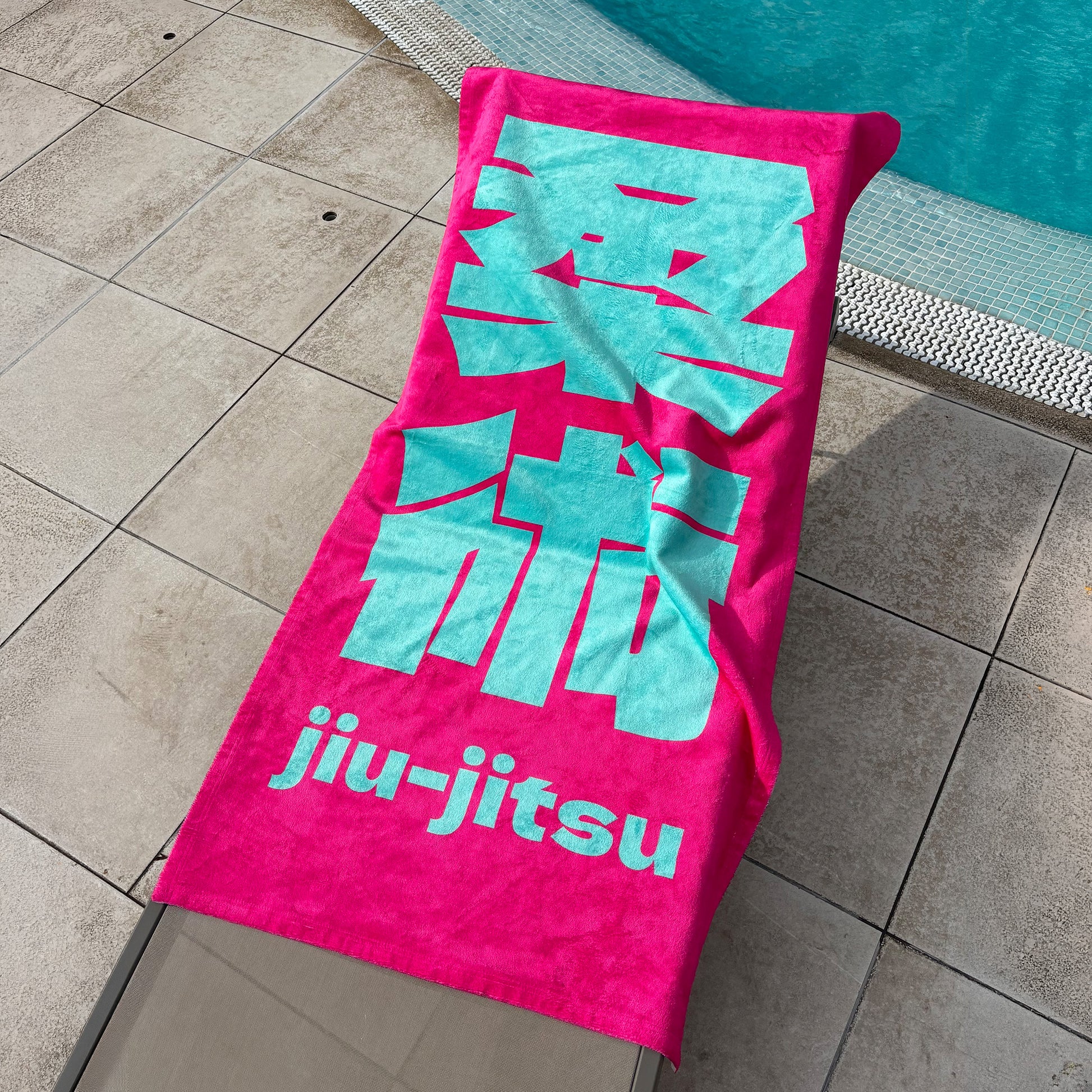 A bright pink towel with large light blue-green Japanese characters and the words 'jiu-jitsu' laid out on a tiled poolside deck.